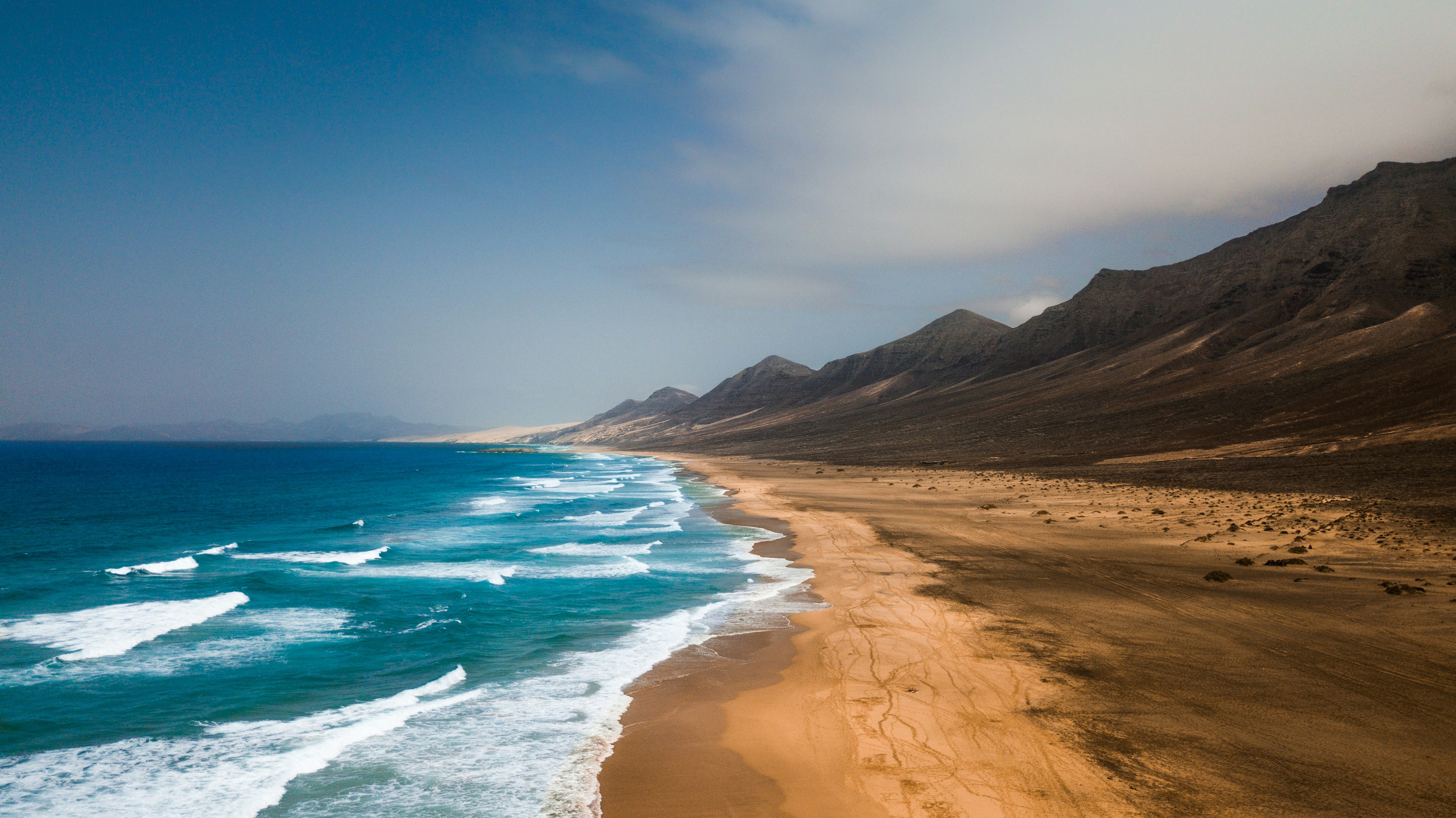 Aerial view of the endless Cofete coastline with turquoise Atlantic waves rolling onto dark sand beneath the Jandía mountain range