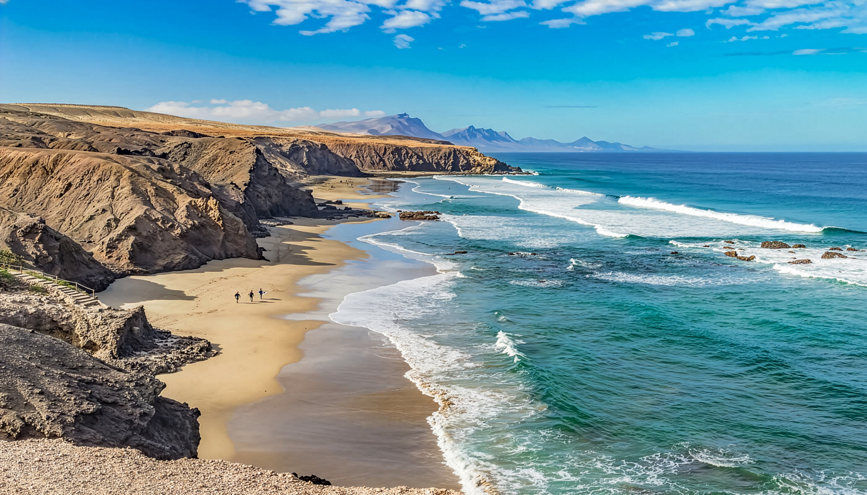 Wide El Cotillo beach framed by rocky cliffs with surfers riding Atlantic waves under blue skies