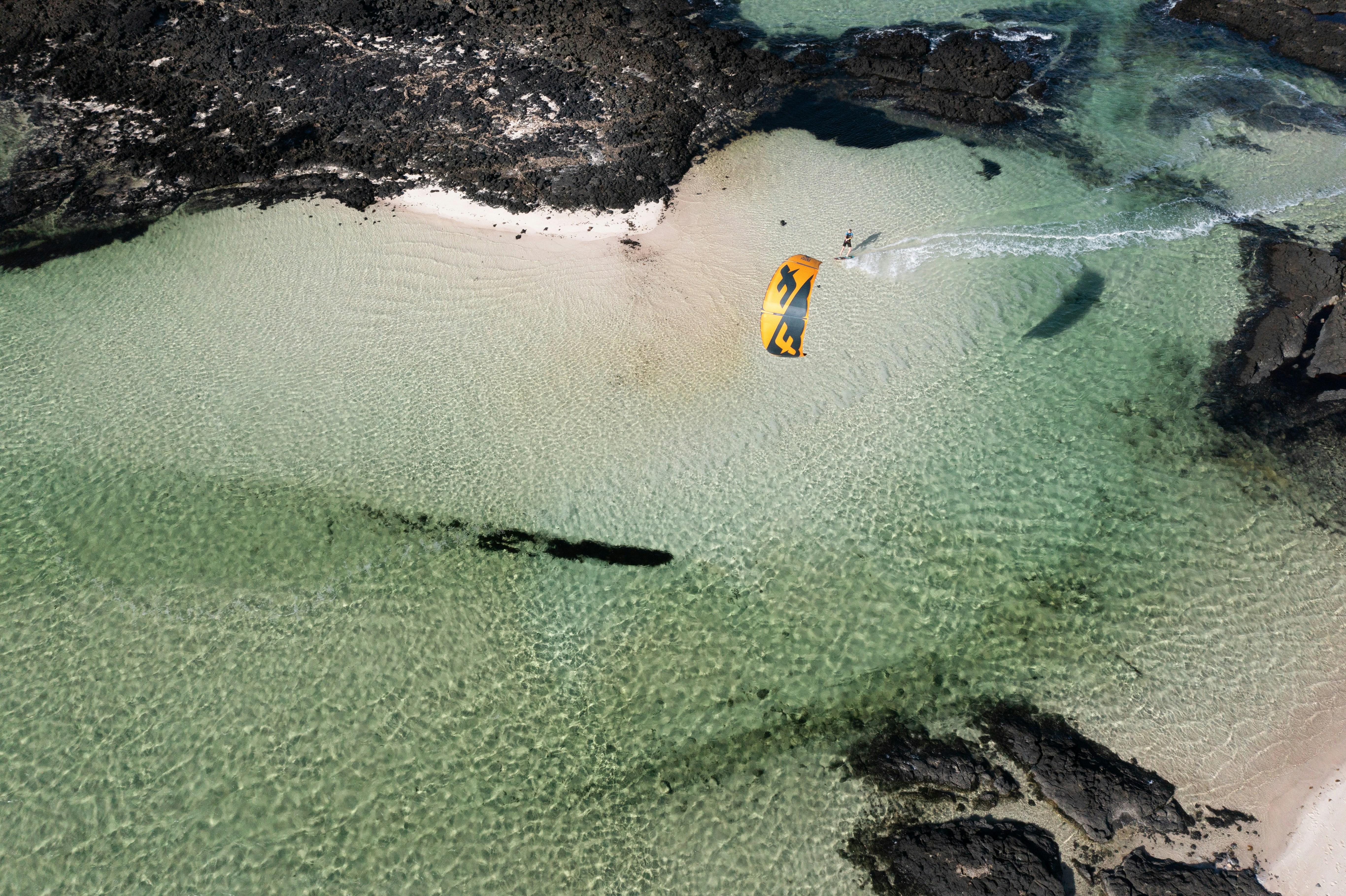 Aerial view of a kite surfer gliding over crystal-clear emerald water in a shallow lagoon sheltered by volcanic rock