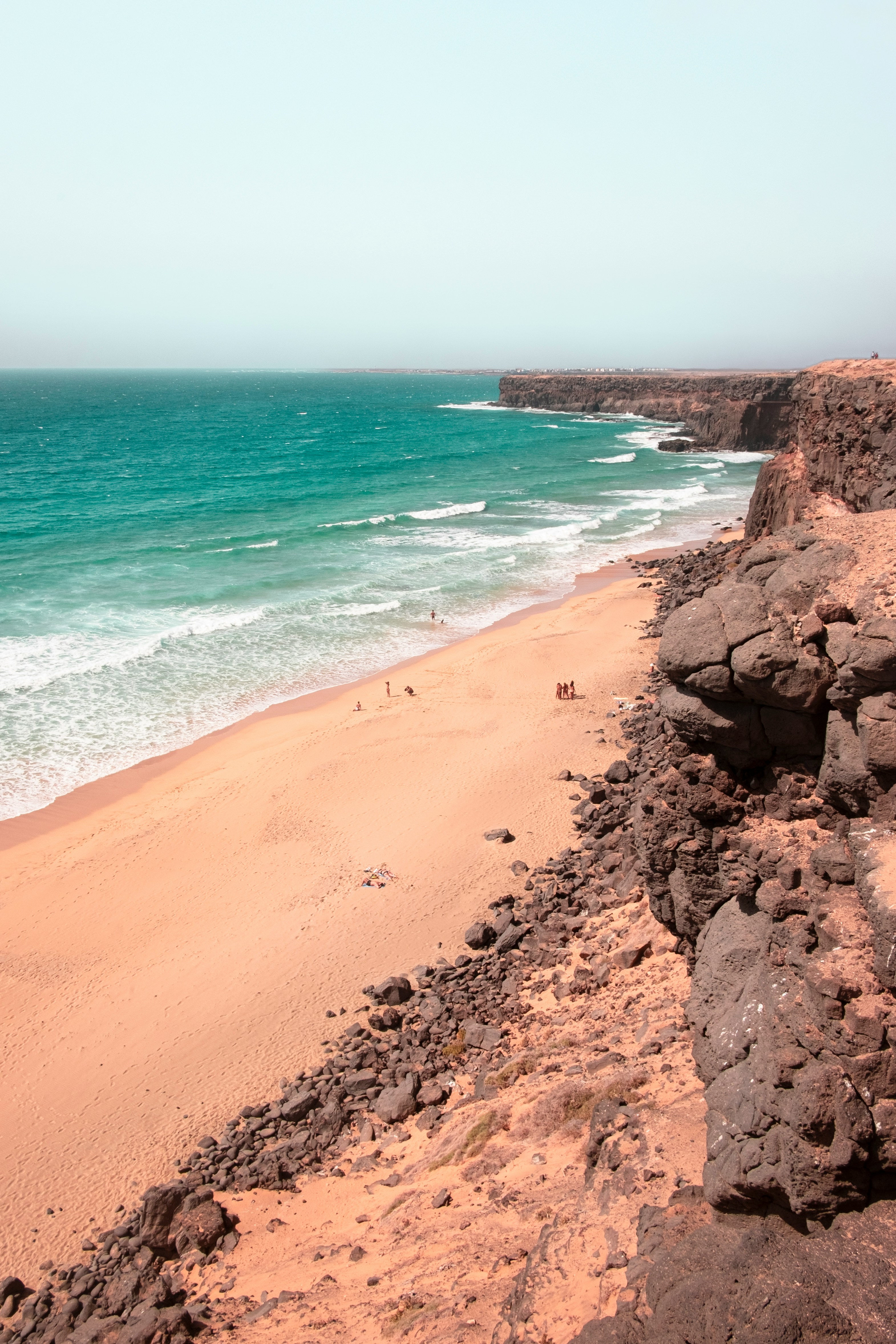 Turquoise waves breaking against rugged west coast cliffs seen from above with sandy beach below